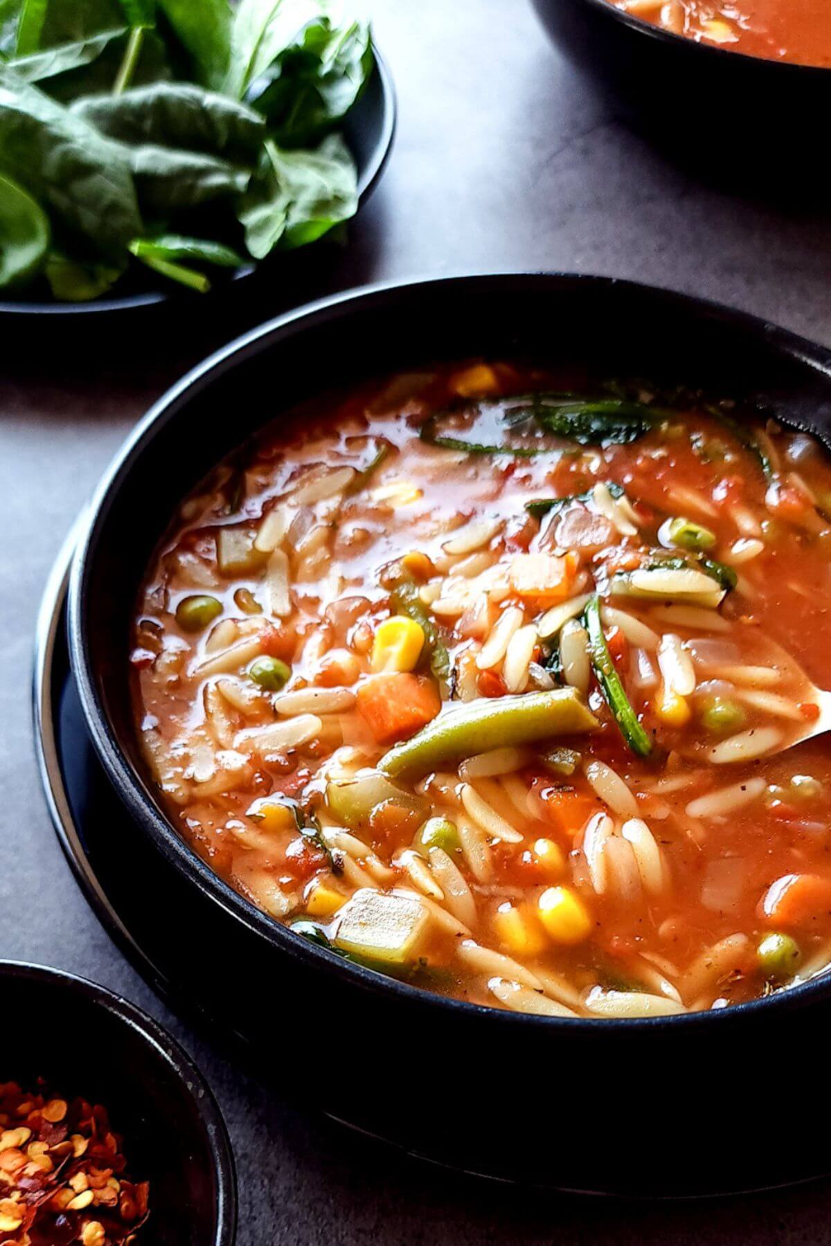 Vegetable orzo soup in a black bowl with bowls of baby spinach and chili flakes on the side.
