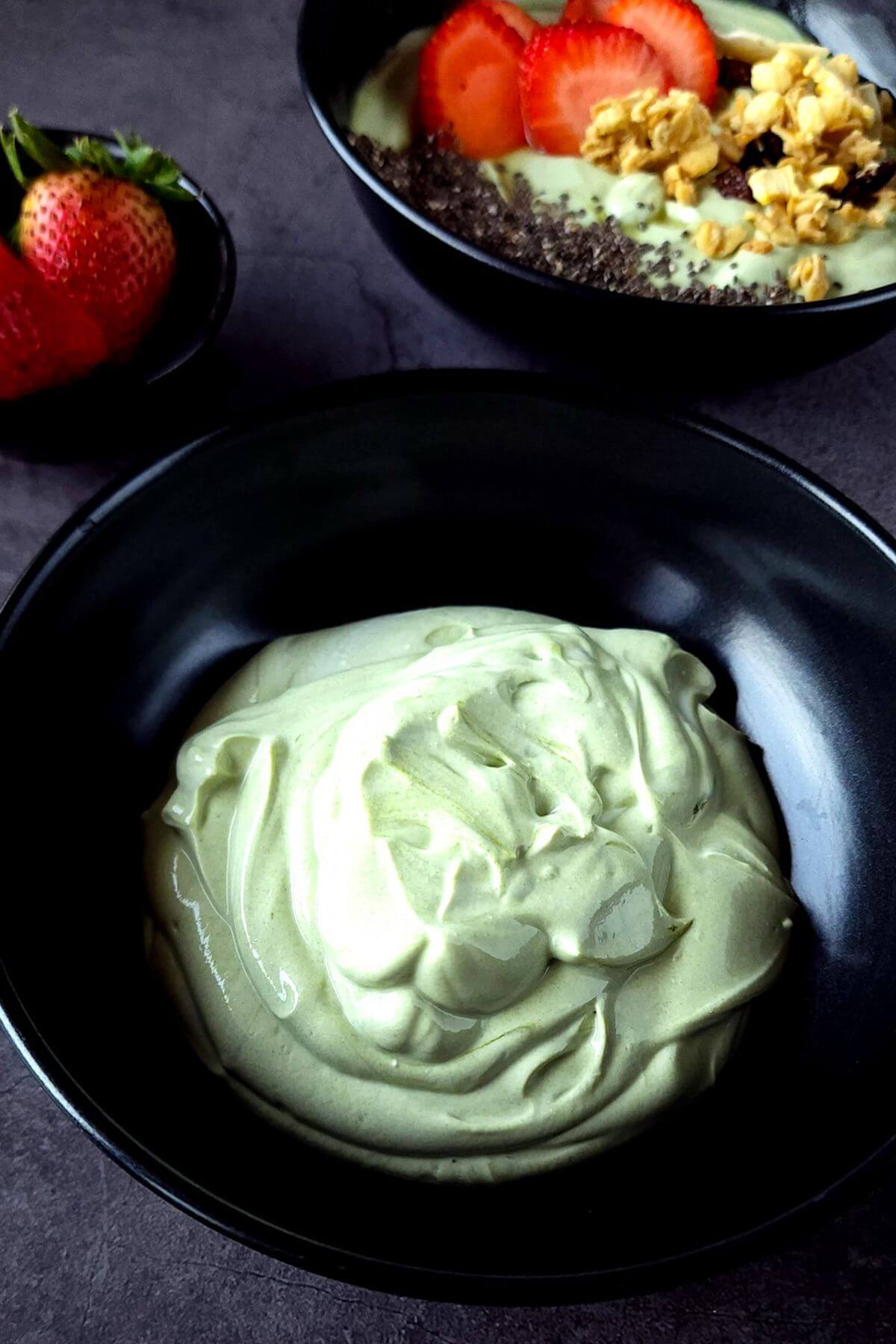Matcha yogurt in a black bowl with a yogurt bowl and a bowl of strawberries in the bcakground.