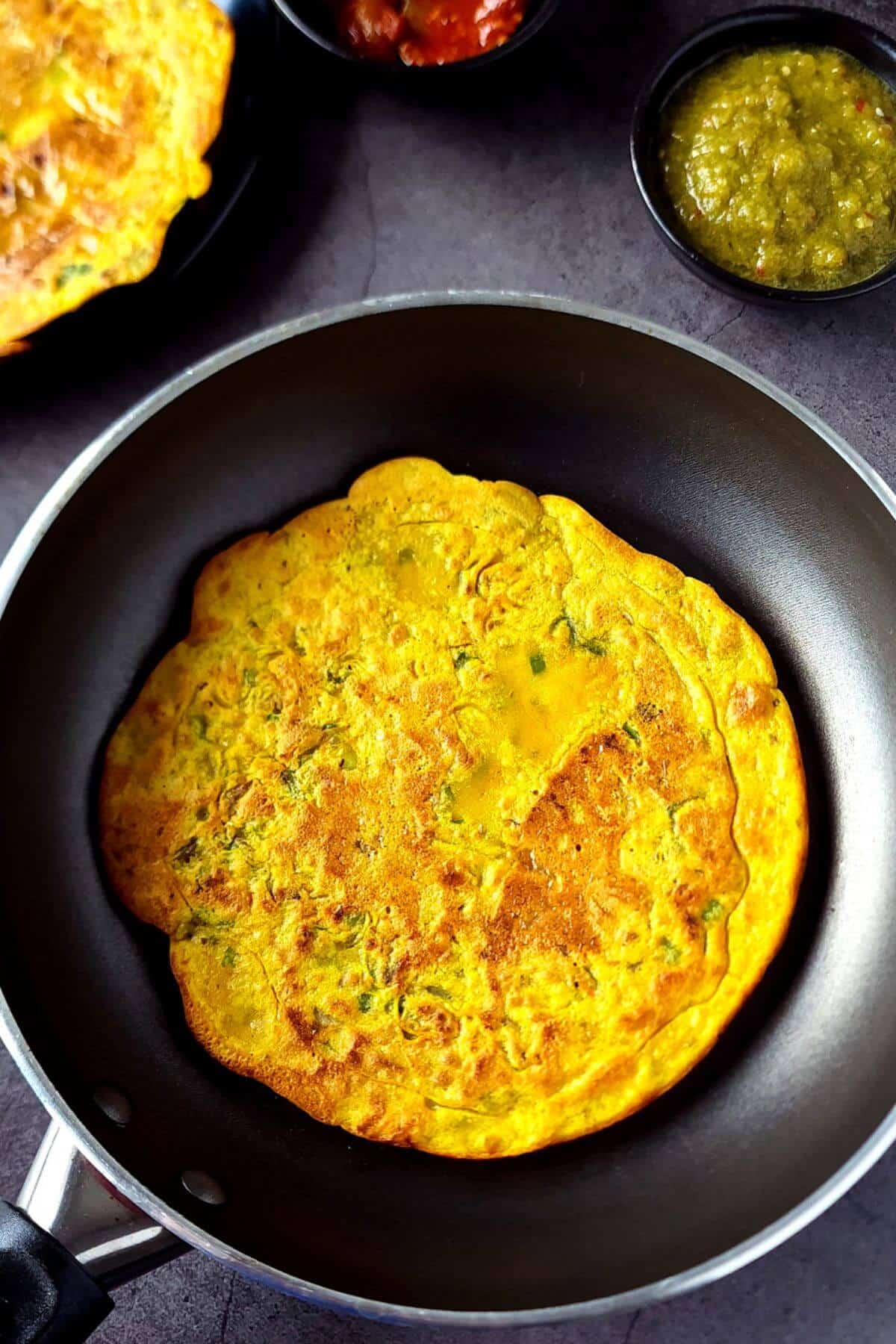 A chickpea flour pancake in a pan with chutneys and pickles on the side