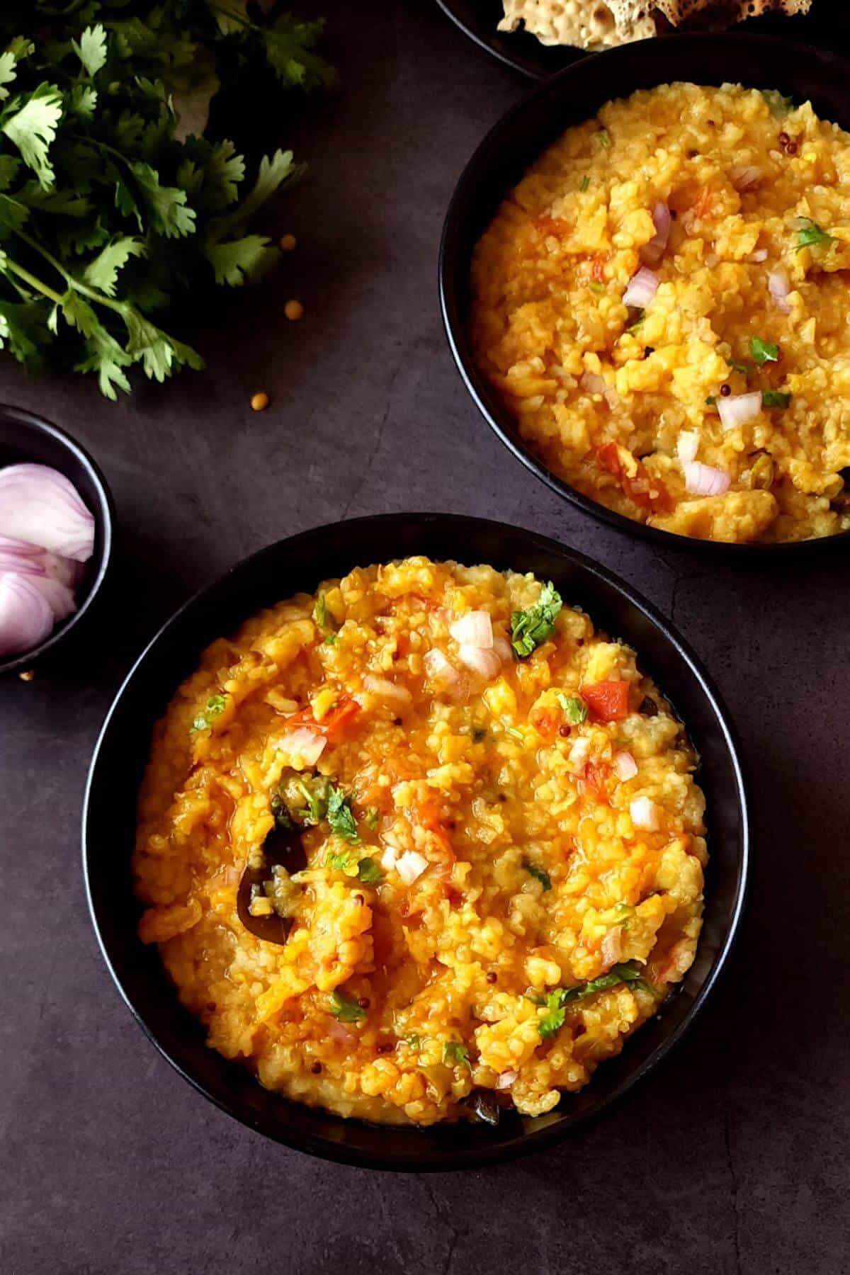 Two bowls of arhar dal khichdi with sliced onion and fresh herbs on the side.