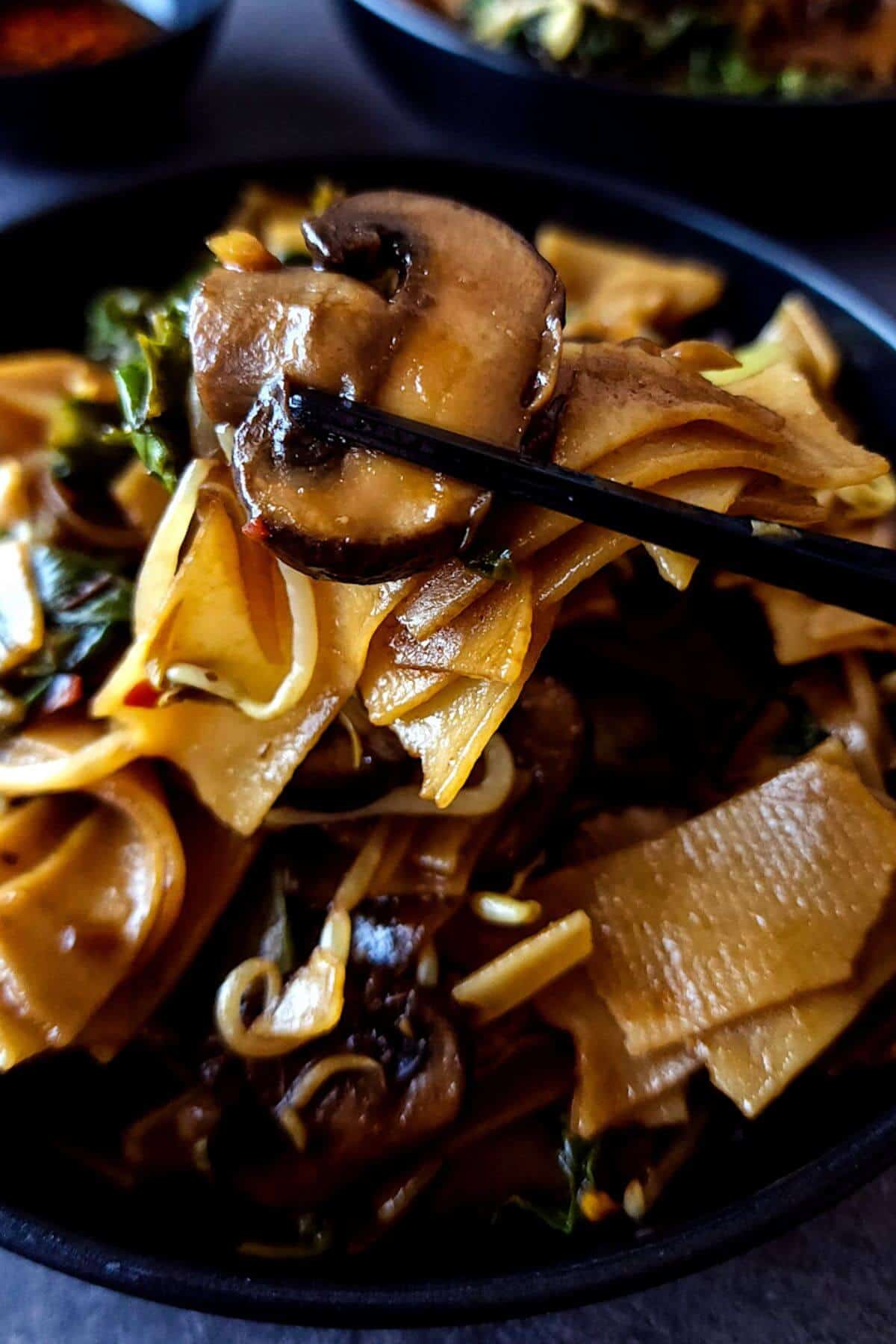 A slice of mushroom picked up with a pair of chopsticks over a bowl of ho fun noodles.