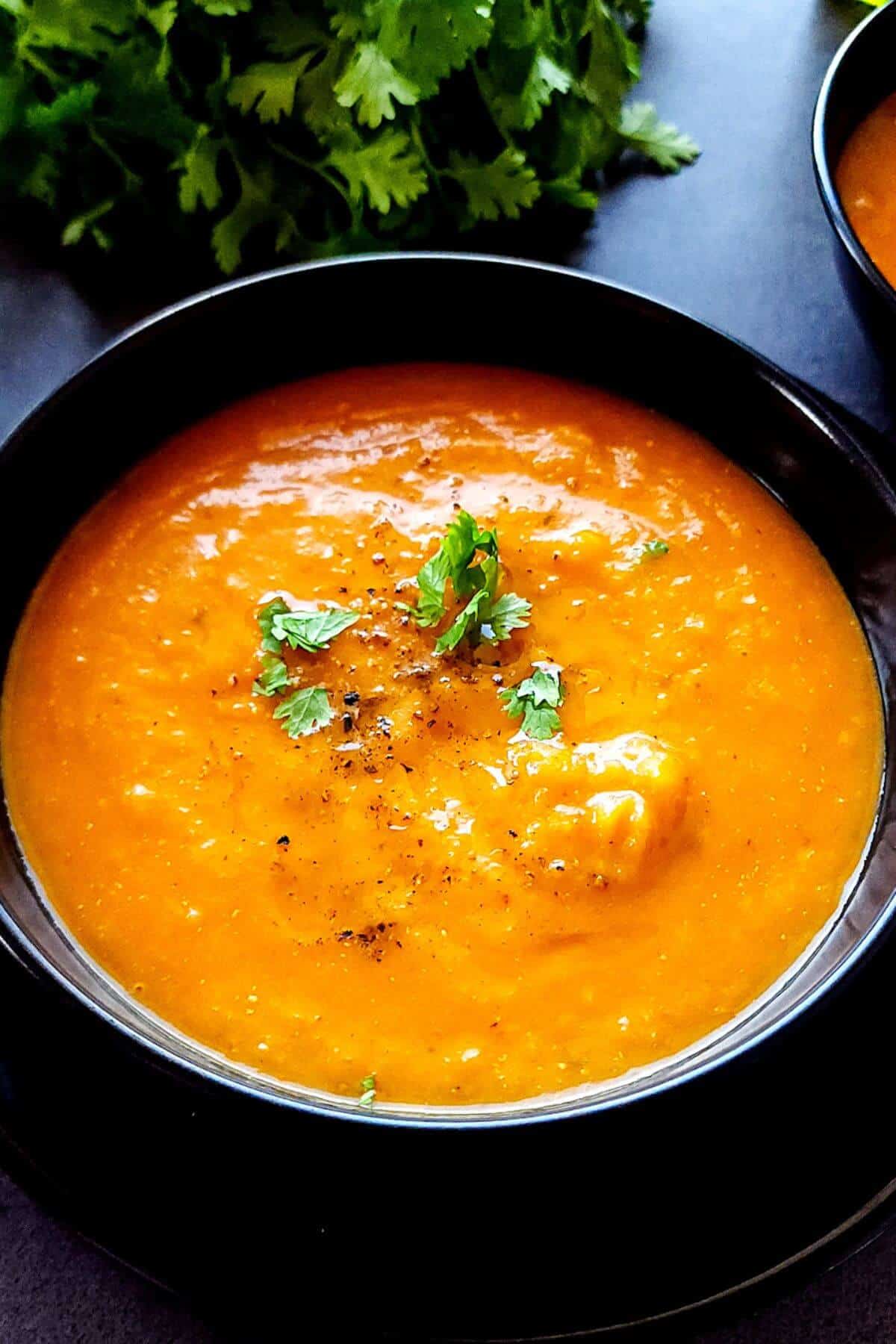 A bowl of butternut squash and red pepper soup with fresh herbs in the background.