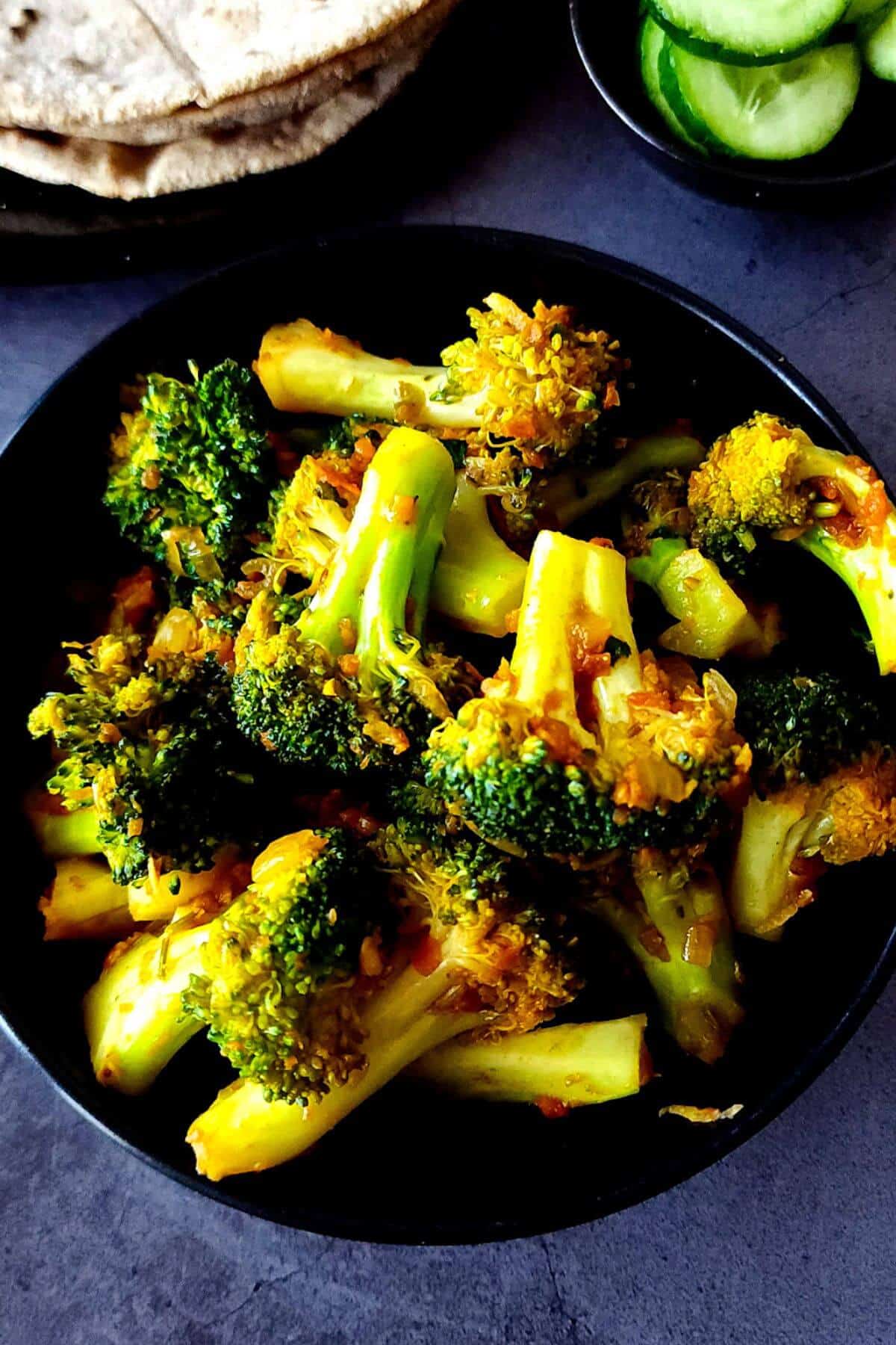 Broccoli curry served in a black bowl with salad and roti in the background.