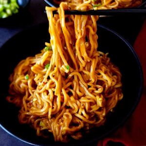 Spicy cheesy ramen lifted up with a pair of chopsticks over a bowl.