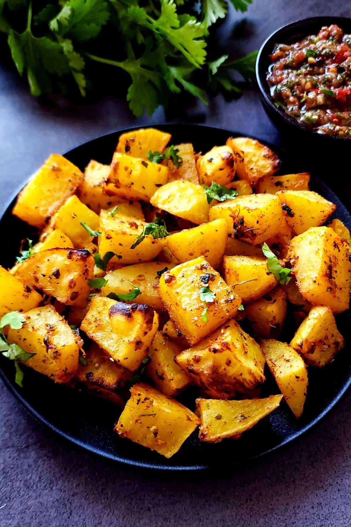A plate of Indian roasted potatoes with a bowl of chutney and fresh herbs in the background.